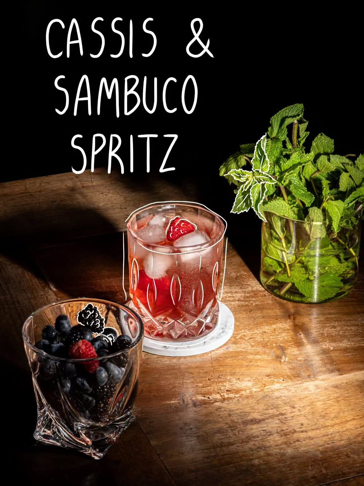 Cassis & Sambuco Spritz in un bicchiere con ghiaccio e frutti rossi, accanto a una ciotola di frutti di bosco e a un vaso di menta
